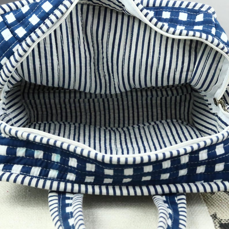 May include: Open view of a blue and white quilted tote bag. The exterior features a checkered pattern, while the interior lining has vertical stripes. The bag has a zipper closure and two handles.