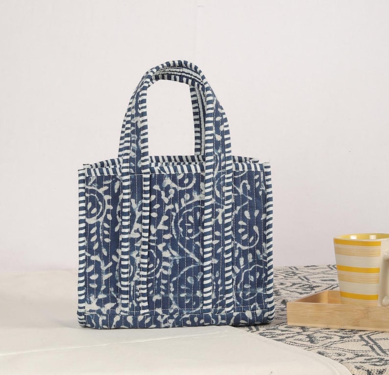 Peut inclure: Un petit sac fourre-tout matelass&eacute; avec un motif floral bleu et blanc. Le sac a deux poign&eacute;es et est fait d'un tissu doux et textur&eacute;. Le sac mesure environ 25 cm de haut et 20 cm de large.