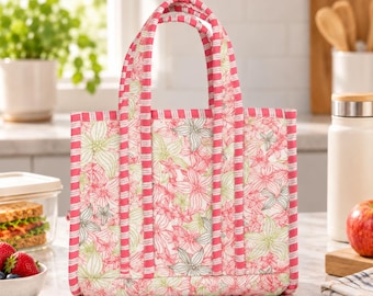 May include: A quilted tote bag featuring a floral pattern in shades of pink, white, and light green. The bag has two vertical handles and a top trim decorated with a pink and white striped fabric pattern, providing a structured and rectangular shape.