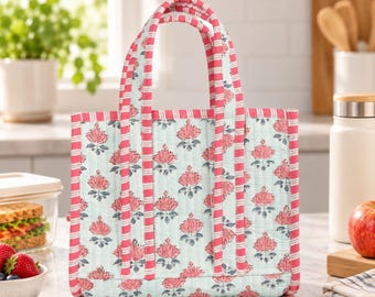 May include: A quilted tote bag featuring a light blue background with a repeating pattern of pink floral motifs. The bag has two handles and trim decorated with pink and white horizontal stripes, providing a structured and functional design.