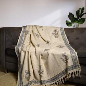 Puede incluir: Una manta de tiro de color crema con un patrón floral azul y marrón. La manta tiene un borde de flecos y está drapeada sobre un sofá gris.