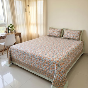 May include: A bed covered in a white cotton sheet featuring a repeating floral pattern in orange and blue, with a matching border. Two rectangular pillows with the same orange and blue floral print and a solid light green trim rest at the head.