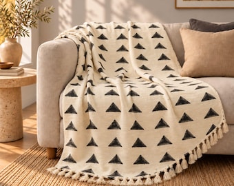 May include: A cream-colored woven throw blanket featuring a repeating pattern of black triangles. The blanket has a textured appearance and is finished with a row of small, matching cream-colored tassels along the bottom edge.