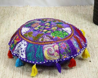 May include: Circular patchwork floor cushion in purple, green, and gold hues. The cushion showcases diverse fabric patterns, including floral and paisley motifs. It's trimmed with colourful tassels and a gold border. A decorative accent for any room, measuring approximately 40cm in diameter.