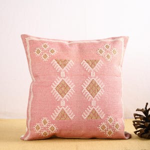 May include: A pink cushion with a geometric pattern in white and gold thread. The cushion is made of a soft, woven fabric.