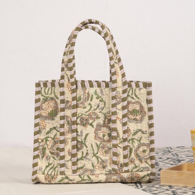 Peut inclure: Un sac fourre-tout matelass&eacute; beige avec un motif floral vert et marron. Le sac a deux anses et une forme rectangulaire. Les bords et les poign&eacute;es sont orn&eacute;s d'un motif &agrave; rayures.