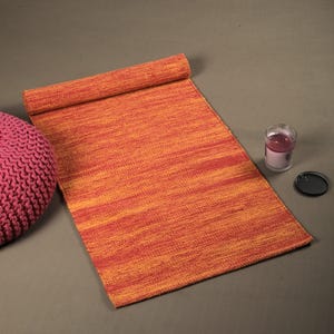 Puede incluir: Una alfombra tejida naranja y roja enrollada con un puf de punto rosa en primer plano.