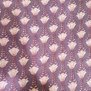 May include: Close-up of a fabric with a repeating floral pattern. The design features white flowers with red accents, arranged vertically on a muted purple background. The fabric has a soft, textured appearance, suitable for various textile projects.