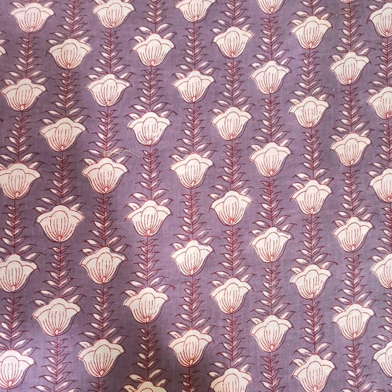 May include: Close-up of a fabric with a repeating floral pattern. The design features white flowers with red accents, arranged vertically on a muted purple background. The fabric has a soft, textured appearance, suitable for various textile projects.