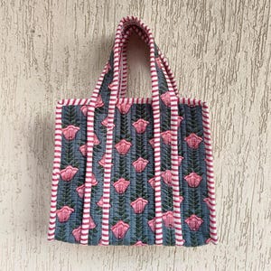 May include: A quilted tote bag with a floral pattern. The bag features a blue background with pink flowers and green leaves. The edges and handles are trimmed with pink and white stripes. The bag is designed for carrying items.