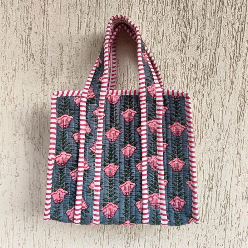 May include: A quilted tote bag with a floral pattern. The bag features a blue background with pink flowers and green leaves. The edges and handles are trimmed with pink and white stripes. The bag is designed for carrying items.