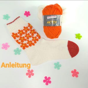 Puede incluir: Un calcetín tejido blanco y naranja con talón y puño rojos, con un estampado floral naranja. También hay un ovillo de lana naranja y recortes de flores decorativas. El texto "Anleitung" es visible.
