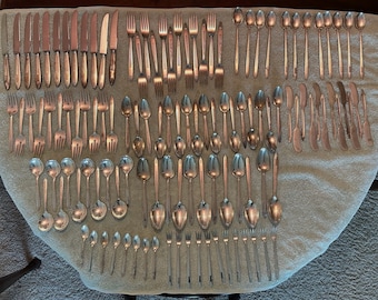 May include: A set of silverware laid out on a white towel. The set includes forks, spoons, and knives. The silverware is arranged in rows, with the forks on the left, the spoons in the middle, and the knives on the right.