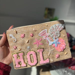 May include: A beige straw clutch bag with the word "HOL" in pink, decorated with starfish, seashells, and a cocktail graphic. The bag has a zip closure and a wrist strap.