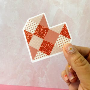 May include: A heart-shaped sticker with a patchwork design. The sticker features various shades of red and pink, with patterns including small hearts and polka dots. The sticker is held against a pink background.