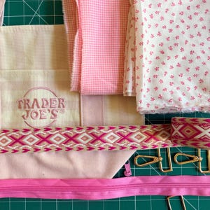 Könnte beinhalten: Eine beige Tragetasche aus Segeltuch mit dem pink gestickten Trader Joe's Logo, begleitet von pinkem Vichy-Stoff, weißem Stoff mit kleinem pinkem Blumenmuster, einem gewebten Band mit pink-weißem Muster, einem pinken Reißverschluss und goldfarbenen Metallteilen.