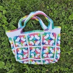 May include: A handmade crochet tote bag with a granny square design. The bag features a white base with colorful squares in shades of pink, blue, yellow, and green. The handles and edges are trimmed with rainbow colors.