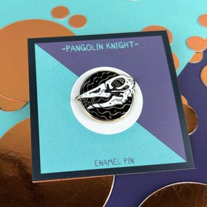 Pode incluir: Pin de esmalte com um design de pangolim a preto e branco, colocado num círculo branco. O pin está num cartão com o texto "-PANGOLIN KNIGHT-" e "ENAMEL PIN". O fundo é turquesa e roxo com círculos cor de cobre.