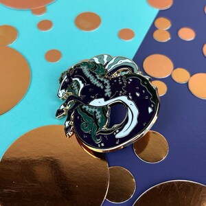May include: A circular enamel pin with a seahorse design in navy blue, white, and green, with gold-coloured metal accents. The pin is set against a backdrop of teal and navy blue, with gold foil circles.