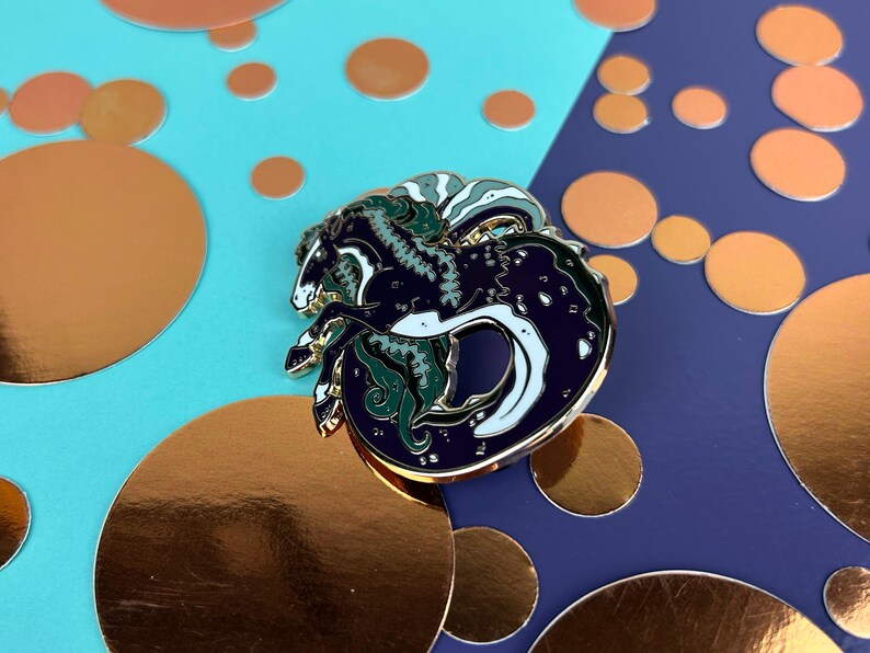May include: A circular enamel pin with a seahorse design in navy blue, white, and green, with gold-coloured metal accents. The pin is set against a backdrop of teal and navy blue, with gold foil circles.