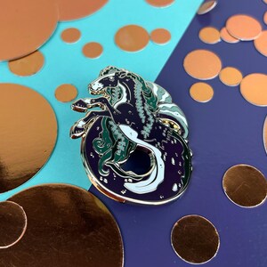 May include: An enamel pin depicting a seahorse, with a dark blue body, white highlights, and green details. The pin is set against a backdrop of copper-coloured circles and a blue and purple background.