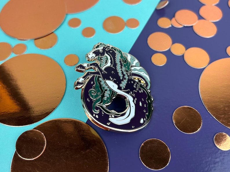 May include: An enamel pin depicting a seahorse, with a dark blue body, white highlights, and green details. The pin is set against a backdrop of copper-coloured circles and a blue and purple background.