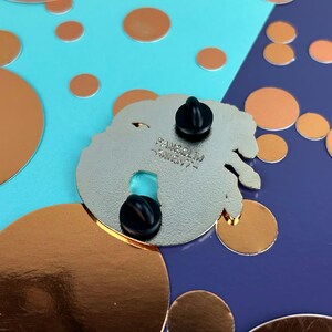 May include: A silver enamel pin with two black pin backs. The pin has a unique shape with a turquoise accent. The text "FANGSOLIM - KNIGHT-" is printed on the back of the pin. The background is a mix of blue and purple with copper-coloured circles.