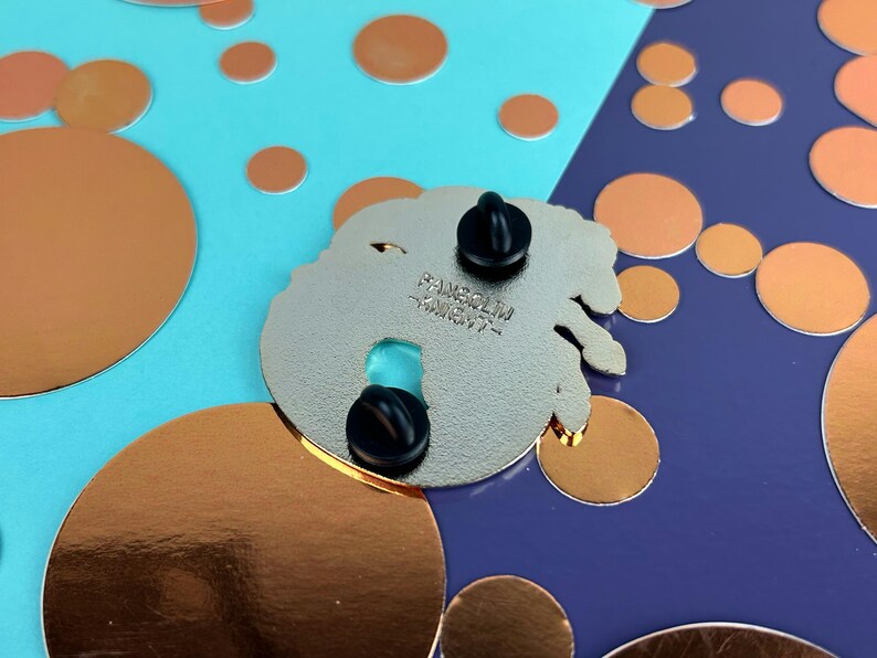 May include: A silver enamel pin with two black pin backs. The pin has a unique shape with a turquoise accent. The text "FANGSOLIM - KNIGHT-" is printed on the back of the pin. The background is a mix of blue and purple with copper-coloured circles.