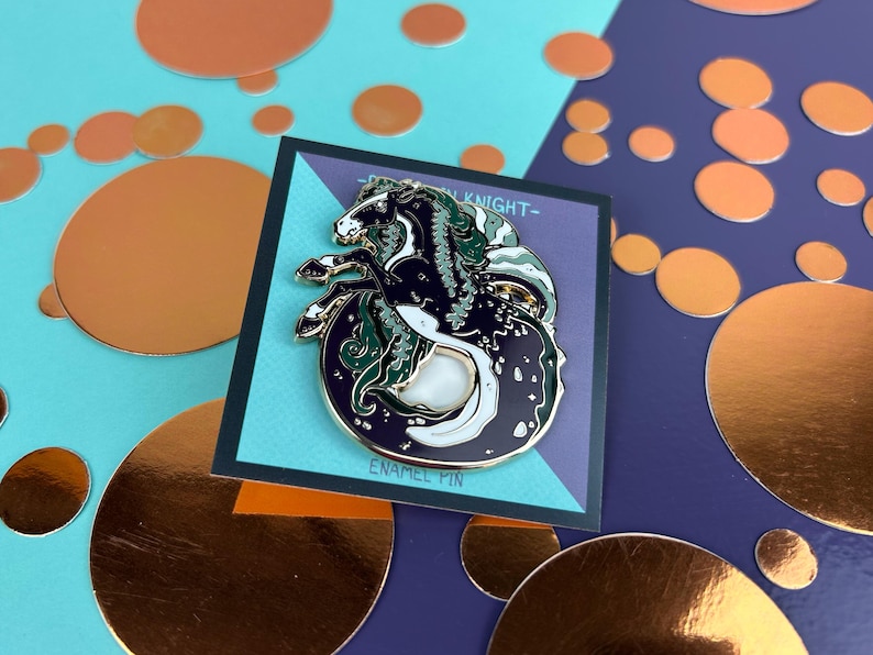 May include: An enamel pin depicting a black and white seahorse with green detailing, set against a dark backdrop. The pin is on a teal and purple card with the text "ENAMEL PIN". The background features copper-coloured circles.