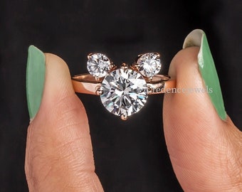 Puede incluir: Un anillo de oro rosa con un gran diamante redondo central y dos diamantes más pequeños a cada lado, con forma de orejas de Mickey Mouse.