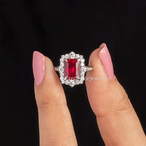 May include: A silver-toned ring featuring a rectangular, deep red center stone surrounded by a halo of small, clear, sparkling stones. The ring has a decorative, scalloped metal frame around the central setting, creating a distinct, ornate appearance.