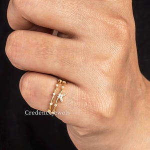 Può includere: Un anello dorato su un dito. L'anello presenta un design delicato con piccole pietre scintillanti e un accento a forma di stella. L'anello è indossato su un dito su sfondo nero.