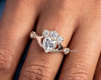 May include: A rose gold ring with a large heart-shaped center stone and smaller stones on the band. The ring is a Celtic knot design.