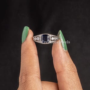 May include: A silver ring featuring a rectangular, dark blue gemstone at the center, flanked by smaller, round, clear stones. The ring has a delicate, ornate design. The ring is held up against a black background.