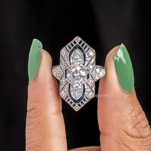 May include: A silver ring with a large, clear center stone surrounded by smaller clear stones and blue stones. The ring is set in a geometric design.