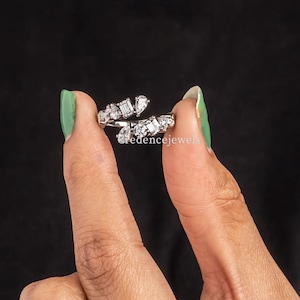 Può includere: Un anello argentato con più pietre chiare, rettangolari e quadrate. L'anello è tenuto tra due dita su uno sfondo nero. L'anello ha il testo "credencejewels".