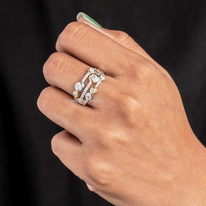 Puede incluir: Un anillo multibanda con un diseño único. El anillo presenta una serie de piedras preciosas redondas y claras engastadas en una banda de plata y oro. Las piedras preciosas varían en tamaño y están dispuestas en un patrón disperso. El anillo se lleva en una mano.