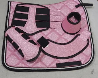 May include: A pink glittery horse saddle pad with a diamond-quilted pattern, black trim, and white piping. The set includes matching pink glittery leg wraps, bell boots, and a curved strap, all featuring black hook-and-loop fasteners.