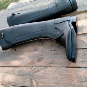 May include: A pair of black tall riding boots featuring a full-length side zipper, a small buckle strap at the top, and a low block heel. The boots have a smooth finish with reinforced stitching along the ankle and calf area for structure.
