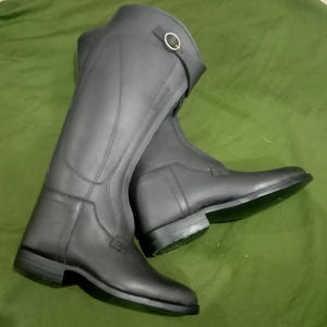 May include: A pair of tall black riding boots featuring a smooth matte finish, structured panels, and a decorative strap near the top opening secured with a small gold-toned metal buckle. The boots have a low heel and a sleek, streamlined design.