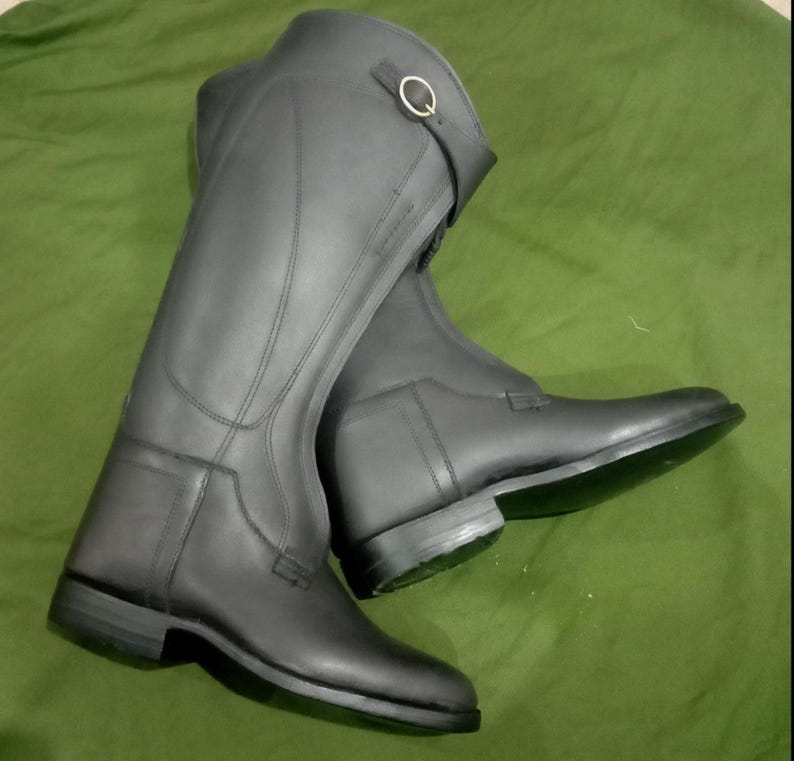 May include: A pair of tall black riding boots featuring a smooth matte finish, structured panels, and a decorative strap near the top opening secured with a small gold-toned metal buckle. The boots have a low heel and a sleek, streamlined design.