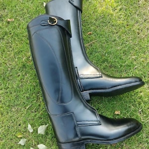 May include: A pair of tall black leather riding boots featuring a smooth finish, a structured shaft, and a decorative strap near the top with a small gold-toned metal buckle. The boots have reinforced stitching along the sides and a low heel.