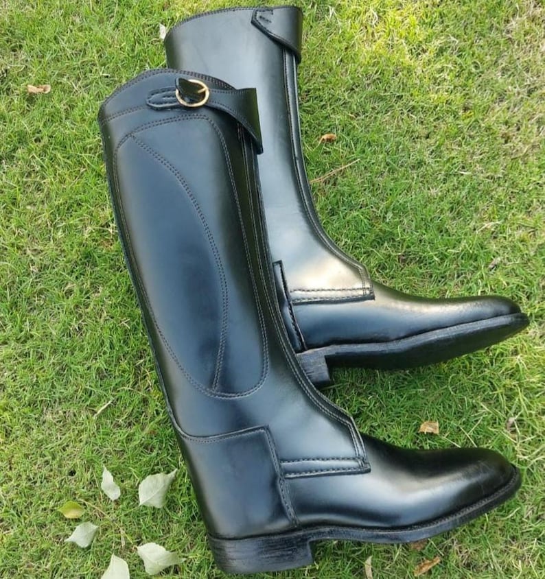 May include: A pair of tall black leather riding boots featuring a smooth finish, a structured shaft, and a decorative strap near the top with a small gold-toned metal buckle. The boots have reinforced stitching along the sides and a low heel.