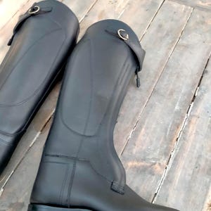 May include: A pair of tall black equestrian riding boots featuring a smooth finish, a structured shaft, and a decorative strap near the top with a metallic buckle closure. The boots have reinforced stitching along the sides and a flat sole design.