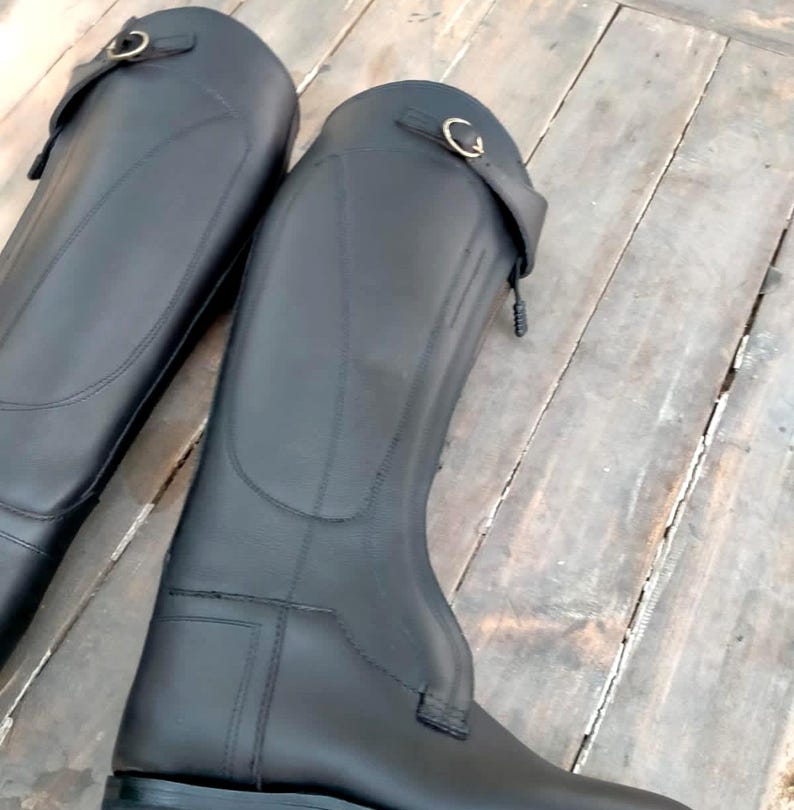 May include: A pair of tall black equestrian riding boots featuring a smooth finish, a structured shaft, and a decorative strap near the top with a metallic buckle closure. The boots have reinforced stitching along the sides and a flat sole design.