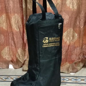 May include: A black boot bag with two top handles and a zippered front pocket. The bag features the gold-colored text Broad Polo Wear, the website www.bwmfg.com, and the social media handle @broadpolowear printed on the front surface.