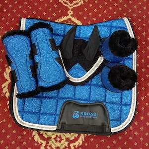 Puede incluir: Un conjunto ecuestre azul con acabado brillante, que incluye una almohadilla para la silla de montar, vendas para las piernas y una cofia. El conjunto presenta detalles y ribetes negros, con el logotipo "BROAD WEAR EQUESTRIAN".
