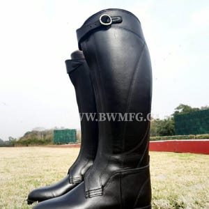 May include: A pair of tall black leather riding boots featuring a structured shaft, a rounded toe, and a decorative strap with a metal buckle near the top. The boots display visible stitching along the sides and the text WWW.BWMFG.COM across the center.