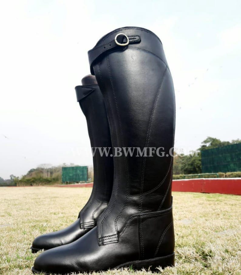 May include: A pair of tall black leather riding boots featuring a structured shaft, a rounded toe, and a decorative strap with a metal buckle near the top. The boots display visible stitching along the sides and the text WWW.BWMFG.COM across the center.