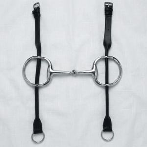 Op de afbeelding: Een zilverkleurig metalen paardenbit met een gebroken mondstuk en twee grote ronde ringen. Twee lange, dunne zwarte leren riemen zijn aan de ringen bevestigd, elk eindigend met een kleine metalen gesp aan de bovenkant en een ring onderaan.